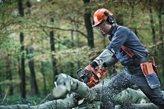 A person using a chainsaw to cut a fallen tree in a forest, wearing work gear and helmet.