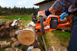 A person using a chainsaw to cut a log outdoors, with wood logs stacked nearby.
