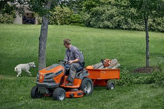 A person driving a ride-on mower with a trailer carrying logs in a garden, with a dog nearby.