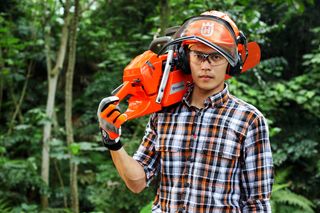 A person carrying a Husqvarna chainsaw on their shoulder in a forest setting.