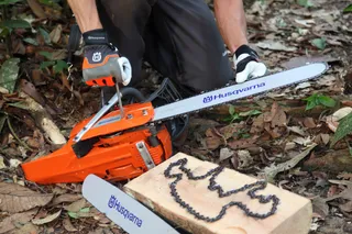 Osoba kleči u šumskom okruženju, podešavajući motornu testeru Husqvarna pored drvenog bloka sa lancem motorne testere.