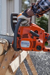 A person using a Husqvarna chainsaw to cut a log on a wooden sawhorse outdoors.