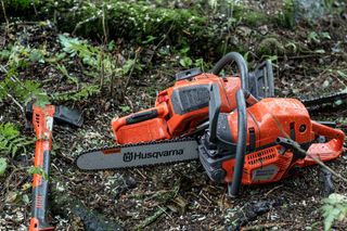 Two Husqvarna chainsaws resting on a forest floor, surrounded by wood chips and foliage.