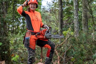 En person i arbetsredskap som håller en motorsåg och står på ett nedfallet träd i en skog.