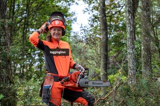 Une personne debout dans une forêt tenant une tronçonneuse Husqvarna, portant des vêtements de travail.