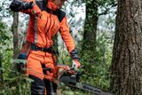 A person in work gear holding a Husqvarna chainsaw in a forest setting.
