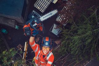 Arborist in work gear climbing a tree with a chainsaw and helmet.