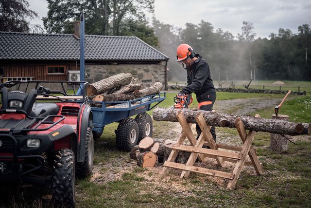 Työvarusteissa oleva henkilö, joka leikkaa moottorisahalla tukkeja sahalla mönkijän ja perävaunun vieressä.