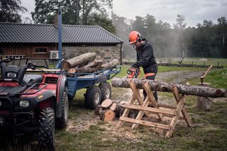 O persoană în echipament de lucru folosind un ferăstrău cu lanț pentru a tăia bușteni pe un fierăstrău lângă un quad și o remorcă.
