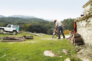 A person using a chainsaw to cut logs in a rural outdoor setting with a vehicle nearby.
