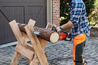 A person using a chainsaw to cut a log on a wooden stand in a garden setting.