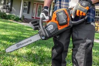 A person holding a Husqvarna chainsaw, showcasing the battery compartment in a garden setting.