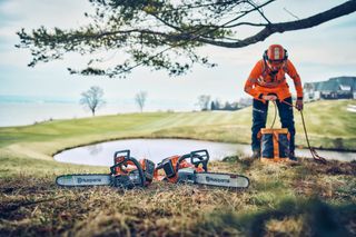Egy munkaruhás személy, aki felszerelést állít fel egy tó közelében, két Husqvarna láncfűrésszel a fűben.