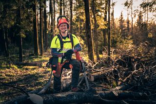 A person in work gear sitting on a fallen tree in a forest, with a chainsaw resting beside them.