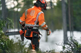 En person i arbetskläder som går i en snöig skog med en motorsåg i handen.