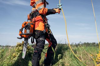 En person iförd arbetsutrustning som håller i ett rep, med en Husqvarna-motorsåg fäst vid sidan, som står i gräsbevuxen terräng.
