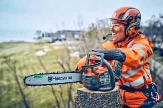 En person i arbetskläder som håller en motorsåg från Husqvarna, med ett suddigt ansikte, på en stubbe.