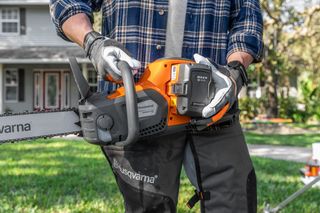A person holding a Husqvarna chainsaw, showcasing the battery compartment in a garden setting.