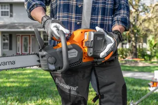 A person holding a Husqvarna chainsaw, showcasing the battery compartment in a garden setting.