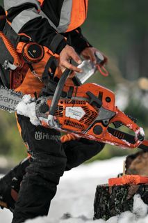 Eine Person in Arbeitskleidung mit einer Husqvarna-Motorsäge, die auf schneebedecktem Boden steht.