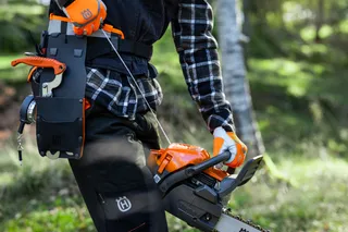 En person i arbetskläder som håller en motorsåg från Husqvarna i skogsmiljö.