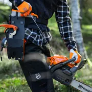 En person i arbetskläder som håller en motorsåg från Husqvarna i skogsmiljö.