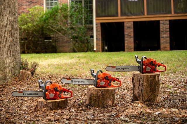 Three Husqvarna chainsaws displayed on tree stumps in a garden setting.