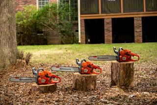 Three Husqvarna chainsaws displayed on tree stumps in a garden setting.
