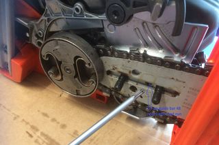 Close-up of a chainsaw's guide bar and chain mechanism with a metal rod pointing at a specific part.