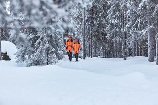 Divi cilvēki, kas staigā pa sniegotu mežu, valkājot spilgtu darba apģērbu.