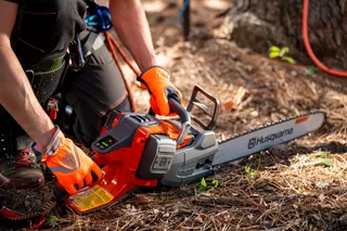 A person kneeling on the ground, inspecting a Husqvarna chainsaw in a forest setting.