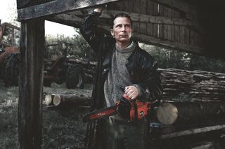 A person holding a chainsaw under a wooden structure, with logs and a tractor in the background.