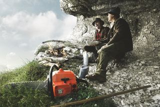 Two people sitting on rocky terrain with a chainsaw and camping gear nearby.