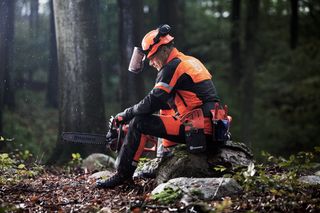 Eine Person in Arbeitskleidung, die auf einem Felsen in einem Wald sitzt und eine Motorsäge in der Hand hält.