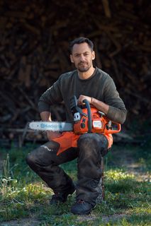 Una persona inginocchiata sull'erba che tiene una motosega Husqvarna davanti a una catasta di legna.