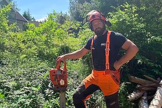 En person som står i skogen og holder en motorsag ved siden av en trestubbe og sager stokker.