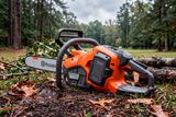Tronçonneuse à batterie Husqvarna sur herbe mouillée, montrant le bloc-batterie à côté des bûches coupées à l’extérieur.