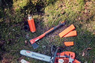 Husqvarna motorzāģis, cirvis un aizsargaprīkojums, kas novietots meža grīdā.