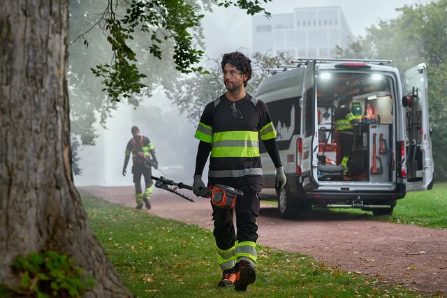 Un trabajador con equipo de trabajo caminando por un camino cerca de un árbol, con una furgoneta y otro trabajador al fondo.