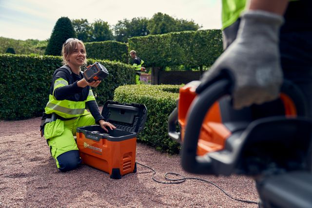 Eine Person, die auf einem Kiesweg kniet und einen Akku in eine Ladestation legt, mit einer Heckenschere im Vordergrund.