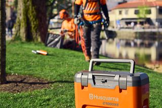 Ящик для зберігання Husqvarna на траві з робітником у верхолазному спорядженні на задньому плані біля дерева.