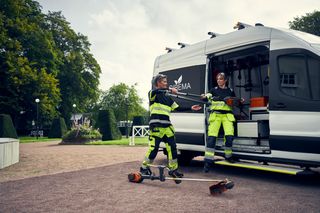 Twee arbeiders in het werktoestel die hulpmiddelen uit een bestelwagen in een tuinomgeving lossen.
