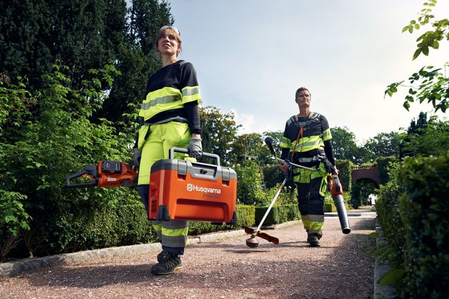 Dvaja pracovníci v záhradnom prostredí, jeden nesie skrinku na náradie Husqvarna a motorovú pílu, druhý drží vyžínač a fúkač lístia.