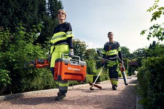 Двоє робітників у саду, один з яких тримає ящик з інструментами Husqvarna та ланцюгову пилку, інший тримає тример і повітродув для листя.