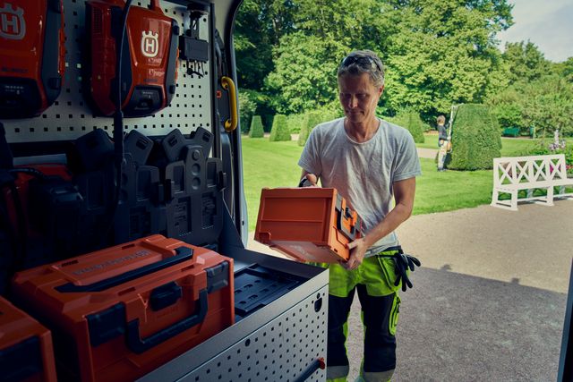 Une personne organise des boîtes à outils Husqvarna dans une fourgonnette garée dans un jardin.