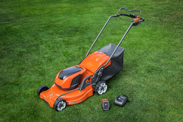 Husqvarnas batteridrivna gräsklippare på en gräsmatta med ett batteri och en laddare bredvid.