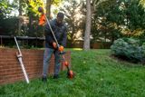 A person using a grass trimmer in a garden setting, with a hedge trimmer resting against a brick wall.