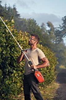 Eine Person, die auf einem Feldweg eine hohe Hecke mit einer langstieligen Heckenschere schneidet.