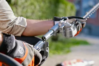 A person using a hedge trimmer attachment on a pole in a garden setting.