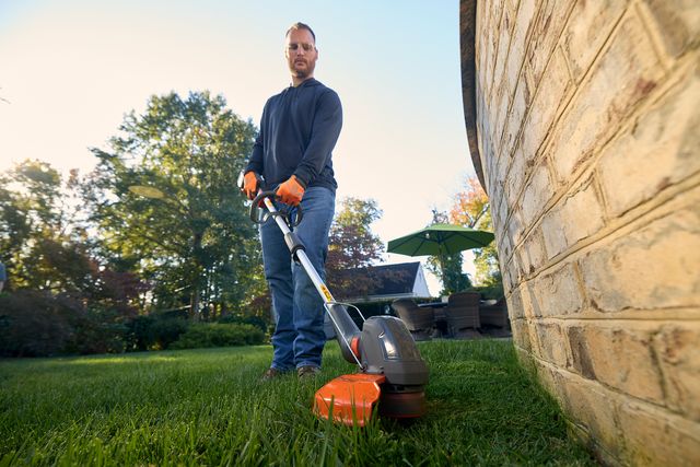 A person using a grass trimmer along the edge of a lawn near a brick wall.